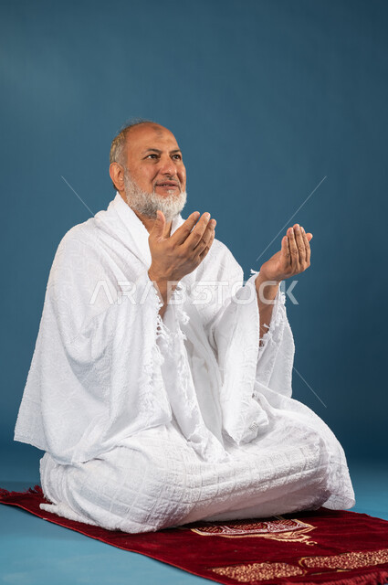 Performing the rituals of Hajj and Umrah, a portrait of an elderly Muslim, Arab, Saudi, and Gulf man wearing ihram clothes and raising his hands up, sitting on the prayer rug, supplication, worship, and closeness to God, the fifth pillar of Islam, Hajj se
