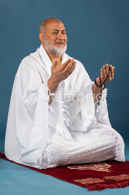 Performing the rituals of Hajj and Umrah, a portrait of an elderly Muslim, Arab, Saudi, and Gulf man wearing Ihram clothes and holding his hand the Rosary and raising his hands up, sitting on the prayer rug, supplication, worship, and closeness to God, th