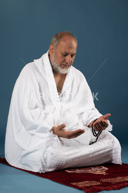 آداء مناسك الحج والعمرة، بورتريه لرجل مسن مسلم عربي سعودي خليجي يرتدي ملابس الاحرام ويمسك بيده السبحة ويرفع يديه لاعلى، الجلوس على سجادة الصلاة والدعاء والعباده والتقرب الى الله، الركن الخامس من أركان الاسلام، موسم الحج 2023، خلفية زرقاء