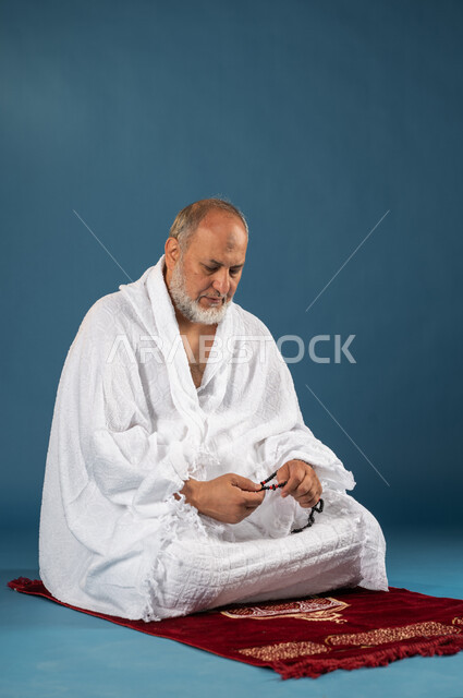 آداء مناسك الحج والعمرة، بورتريه لرجل مسن مسلم عربي سعودي خليجي يرتدي ملابس الاحرام ويمسك بيده السبحة، الجلوس على سجادة الصلاة، التسبيح والاستغفار وذكر الله تعالى، العباده والتقرب الى الله، الركن الخامس من أركان الاسلام، موسم الحج 2023، خلفية زرقاء