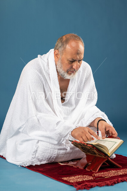 حفظ وتفسير القران الكريم، بورتريه لرجل مسن مسلم خليجي سعودي يجلس على سجادة الصلاة ويقوم بقراءة وتلاوة القران، ارتداء ملابس الاحرام، آداء مناسك الحج والعمرة، الركن الخامس من أركان الاسلام، موسم الحج 2023، خلفية زرقاء