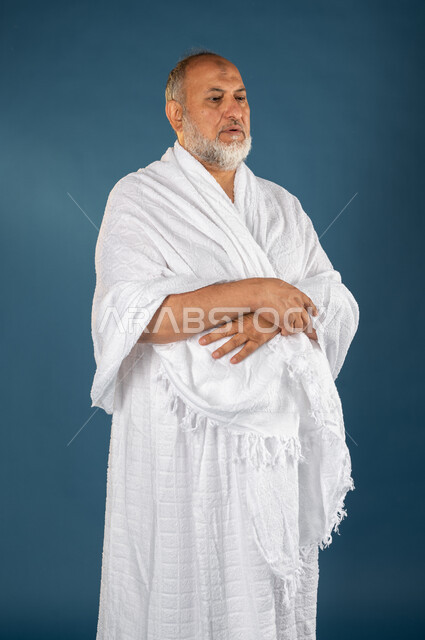 اداء فريضة الصلاة، بورتريه لرجل مسن مسلم خليجي سعودي يرتدي ملابس الاحرام ويجلس على سجادة الصلاة، آداء مناسك الحج والعمرة، التضرع والتقرب إلى الله، الركن الخامس من أركان الاسلام، موسم الحج 2023، خلفية زرقاء