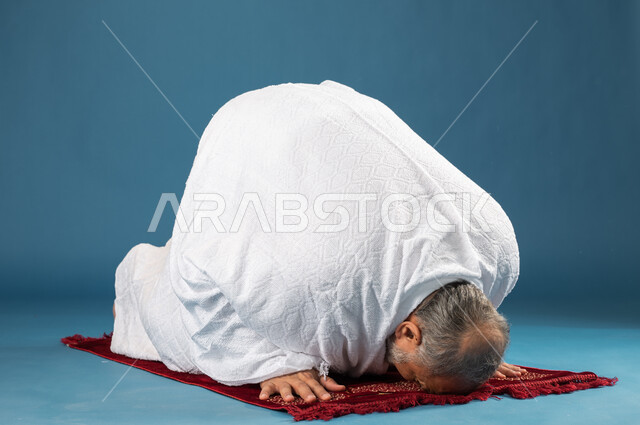 السجود والتضرع والتقرب إلى الله، بورتريه لرجل مسن مسلم خليجي سعودي يرتدي ملابس الاحرام ويجلس على سجادة الصلاة، اداء فريضة الصلاة، آداء مناسك الحج والعمرة، الركن الخامس من أركان الاسلام، موسم الحج 2023، خلفية زرقاء