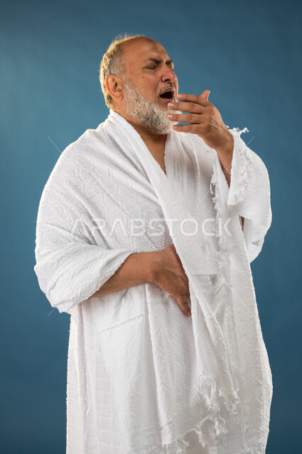 ارتداء ملابس الاحرام، العطاس والانفلونزا، بورتريه لرجل مسن مسلم عربي سعودي خليجي مريض يعطس، إيماءات وجه تدل على المرض والتعب، آداء مناسك الحج والعمرة، الركن الخامس من أركان الاسلام، موسم الحج 2023، خلفية زرقاء