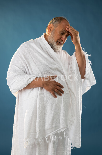 وجع الرأس والصداع، الإحساس بالمرض، بورتريه لرجل مسن مسلم عربي سعودي خليجي يرتدي ملابس الاحرام ويضع يده على رأسه، إيماءات وجه ويدين تدل على الألم والتعب، الركن الخامس من أركان الاسلام، موسم الحج 2023، خلفية زرقاء