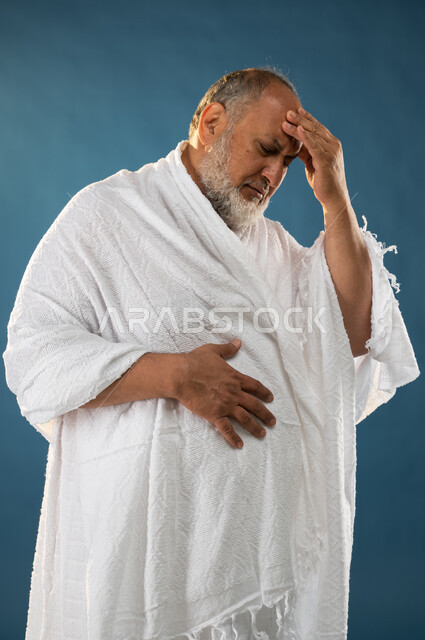 Headache and headache, feeling sick, a portrait of an elderly Muslim Arab Saudi Gulf man wearing ihram clothes and placing his hand on his head, facial and hand gestures indicating pain and fatigue, the fifth pillar of Islam, Hajj season 2023, blue backgr