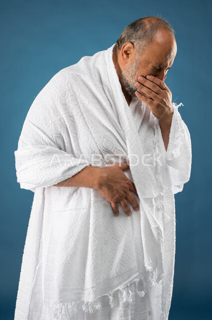 ارتداء ملابس الاحرام، بورتريه لرجل مسن مسلم عربي سعودي خليجي يضع يده على فمه، العطاس والانفلونزا، إيماءات وجه ويدين تدل على المرض والتعب، آداء مناسك الحج والعمرة، الركن الخامس من أركان الاسلام، موسم الحج 2023، خلفية زرقاء