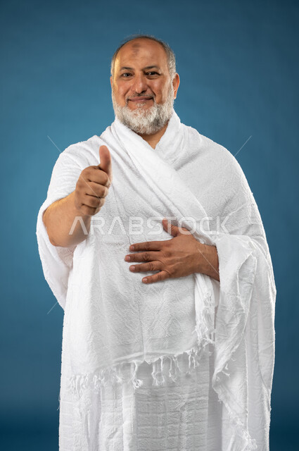 ارتداء ملابس الاحرام، بورتريه لرجل مسن مسلم خليجي سعودي مبتسم يرفع اصبع الابهام لاعلى، إيماءات وجه ويدين تدل على القبول والموافقة والإعجاب، آداء مناسك الحج والعمرة، الركن الخامس من أركان الاسلام، موسم الحج 2023، خلفية زرقاء