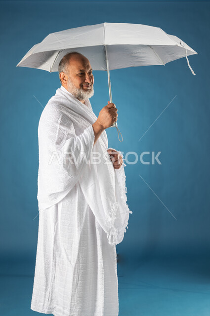 آداء مناسك الحج والعمرة، بورتريه لرجل مسن مسلم خليجي عربي سعودي يرتدي ملابس الاحرام ويحمل بيده شمسية رمادية صغيرة، موسم الحج 2023، الركن الخامس من أركان الاسلام، خلفية زرقاء