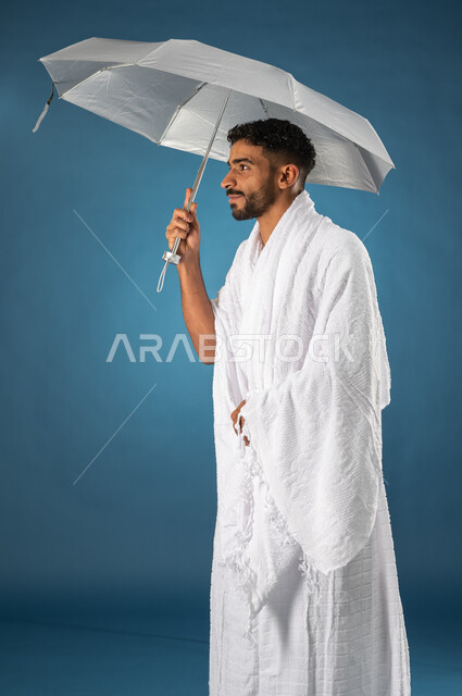 بورتريه لشاب مسلم خليجي عربي سعودي يرتدي ملابس الاحرام ويحمل بيده شمسية رمادية صغيرة، موسم الحج 2023، آداء مناسك الحج والعمرة، الركن الخامس من أركان الاسلام، خلفية زرقاء