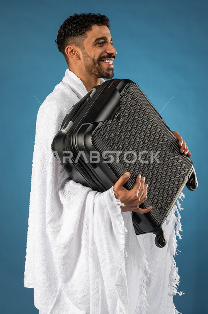 الذهاب لاداء فريضة الحج، بورتريه لشاب مسلم خليجي عربي سعودي يرتدي ملابس الاحرام ويحمل حقيبة السفر، الاستعداد للسفر، ايماءات وجه تدل على السعادة، موسم الحج 2023، آداء مناسك الحج والعمرة، الركن الخامس من أركان الاسلام، خلفية زرقاء