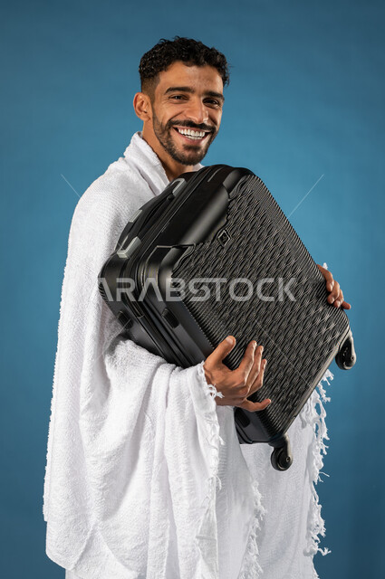 الذهاب لاداء فريضة الحج، بورتريه لشاب مسلم خليجي عربي سعودي يرتدي ملابس الاحرام ويحمل حقيبة السفر، الاستعداد للسفر، ايماءات وجه تدل على السعادة، موسم الحج 2023، آداء مناسك الحج والعمرة، الركن الخامس من أركان الاسلام، خلفية زرقاء