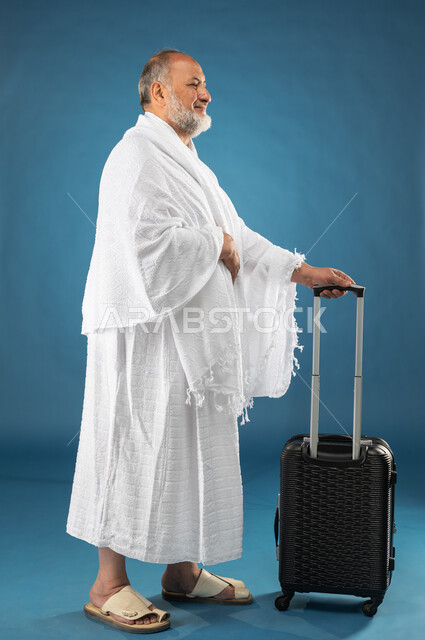الاستعداد للسفر والذهاب لاداء فريضة الحج، بورتريه لرجل مسن مسلم خليجي عربي سعودي يمسك حقيبة السفر ويرتدي ملابس الاحرام، موسم الحج 2023، آداء مناسك الحج والعمرة، الركن الخامس من أركان الاسلام، صورة الجسم بالطول الكامل، خلفية زرقاء