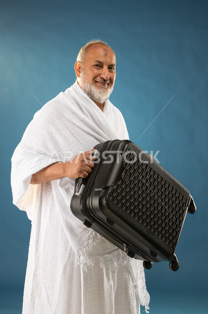 ارتداء ملابس الاحرام، بورتريه لرجل مسن مسلم خليجي عربي سعودي يحمل حقيبة السفر، الاستعداد للسفر والذهاب لاداء فريضة الحج، ايماءات وجه تدل على السعادة، موسم الحج 2023، آداء مناسك الحج والعمرة، الركن الخامس من أركان الاسلام، خلفية زرقاء