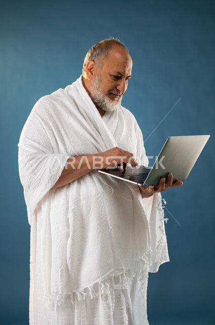 تصفح الانترنت والمواقع الالكترونية، بورتريه لرجل مسن مسلم خليجي عربي سعودي يرتدي ملابس الاحرام ويستخدم الحاسوب المحمول، تصفح مواقع الارشادات لاداء مناسك الحج والعمرة، الركن الخامس من أركان الاسلام، موسم الحج 2023، خلفية زرقاء