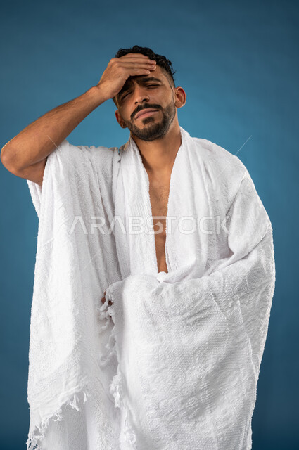وجع الرأس والصداع والإحساس بالمرض، بورتريه لشاب مسلم عربي سعودي خليجي يرتدي ملابس الاحرام ويضع يده على رأسه، إيماءات وجه ويدين تدل على الألم والتعب، الركن الخامس من أركان الاسلام، موسم الحج 2023، خلفية زرقاء