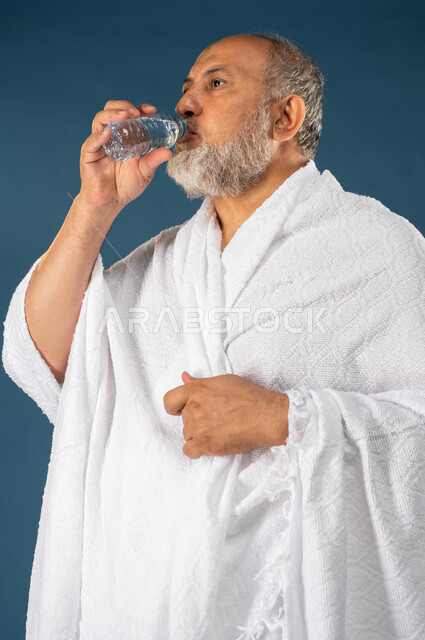 الارتواء بالمياه العذبة، بورتريه لرجل مسن مسلم عربي سعودي خليجي يرتدي ملابس الاحرام ويستمتع بشرب مياه زمزم، اداء مناسك الحج والعمرة، الركن الخامس من أركان الاسلام، موسم الحج 2023، خلفية زرقاء