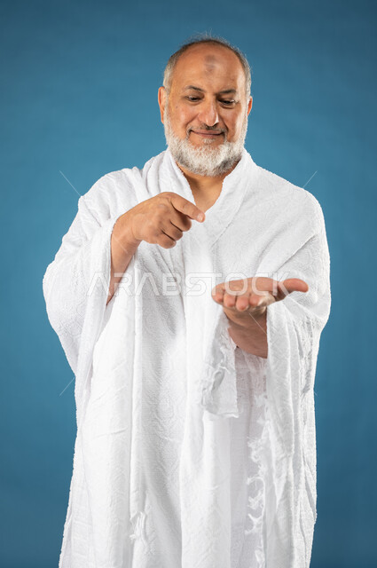 حركة اصبع واشارات، بورتريه لرجل مسن مسلم خليجي سعودي عربي يرتدي ملابس الاحرام ويشير بأصبع السبابة لكف يده الاخرى لعرض شيئاً ما أمامه، آداء مناسك الحج والعمرة، الركن الخامس من أركان الاسلام، موسم الحج 2023، خلفية زرقاء