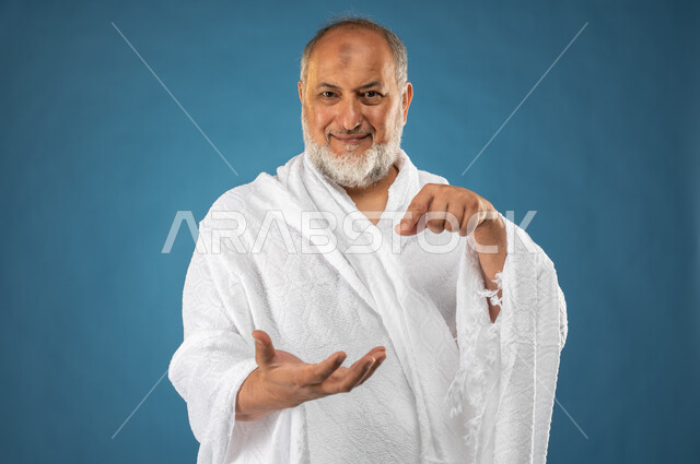 حركة اصبع واشارات، بورتريه لرجل مسن مسلم خليجي سعودي عربي يرتدي ملابس الاحرام ويشير بأصبع السبابة لكف يده الاخرى، حمل ورفع شيء بيديه، عرض شيئاً ما أمامه، آداء مناسك الحج والعمرة، الركن الخامس من أركان الاسلام، موسم الحج 2023، خلفية زرقاء