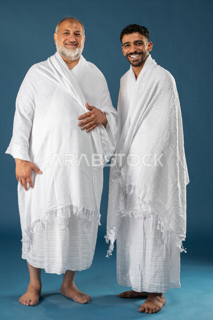 اب مسن وابنه بايماءات وجه تدل على السعادة، بورتريه لرجلان عربيان خليجيان سعوديان يرتديان ملابس الاحرام ويبتسمان، آداء مناسك الحج والعمرة، الركن الخامس من أركان الاسلام، موسم الحج 2023، صورة الجسم بالطول الكامل، خلفية زرقاء