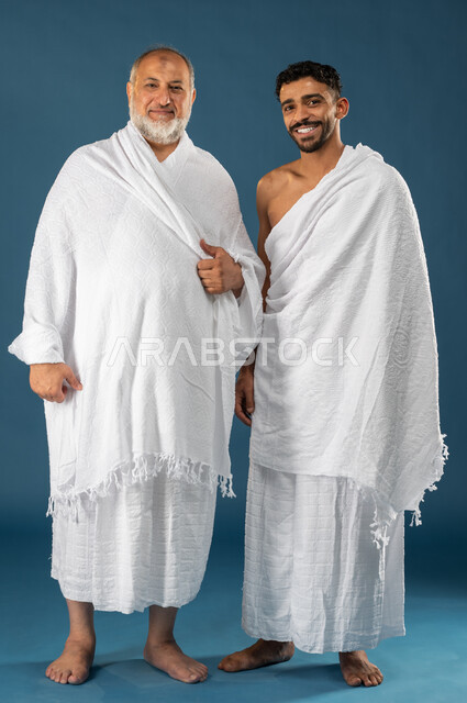 اب مسن وابنه بايماءات وجه تدل على السعادة، بورتريه لرجلان عربيان خليجيان سعوديان يرتديان ملابس الاحرام ويبتسمان، آداء مناسك الحج والعمرة، الركن الخامس من أركان الاسلام، موسم الحج 2023، صورة الجسم بالطول الكامل، خلفية زرقاء
