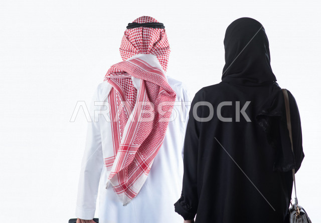 بورتريه من الخلف لزوج عربي خليجي سعودي يمسك بيده حقيبة سفر يقف بجانب زوجته استعداداً لرحلة سياحية ، جولة سياحية ، سياحة و سفر ، قضاء أوقات ممتعة ، خلفية بيضاء