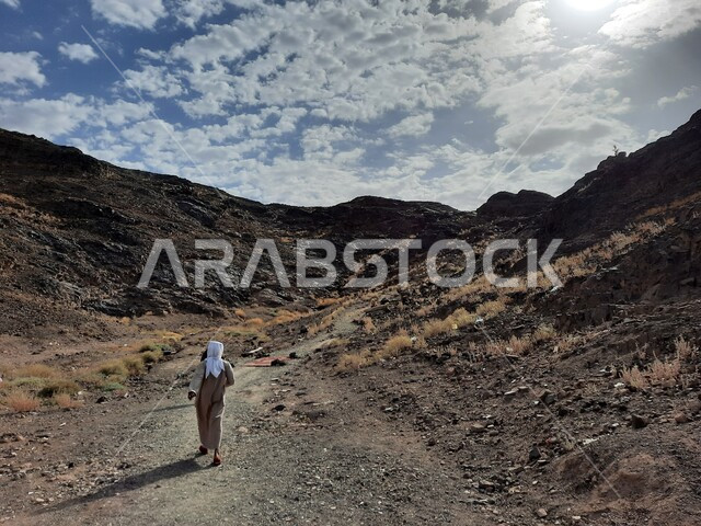هواية تسلق الجبال، صورة من الخلف لرجل عربي خليجي سعودي يتسلق جبال المدينة المنورة في المملكة العربية السعودية، القمم والمرتفعات الجبلية، جبال المدينة المنورة الصخرية، تضاريس الطبيعة الجبلية في السعودية، اماكن سياحية في السعودية