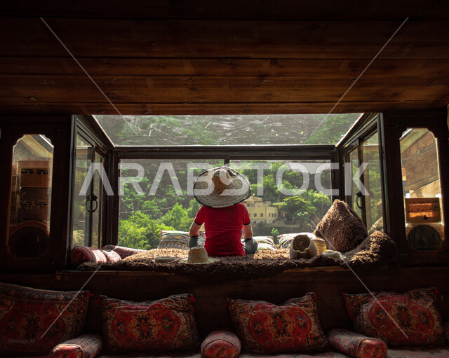 صورة من الخلف لسائح يرتدي قبعة ويجلس امام نافذة كوخ في قرية رجال ألمع، الاشجار والنباتات الخضراء، جلسات واكواخ شعبية في عسير في المملكة العربية السعودية، السياحة في السعودية