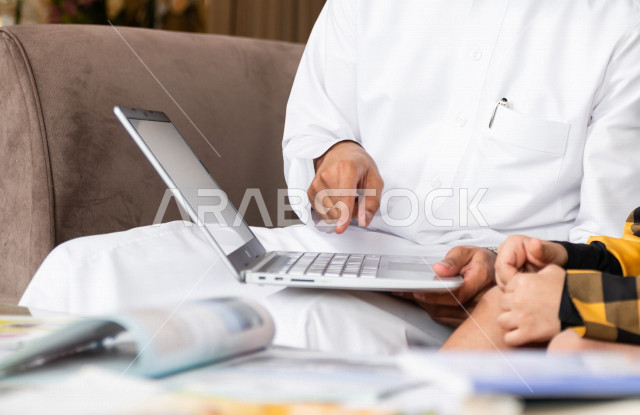 A Saudi Gulf student studying at home with his father, who follows with him online classes via a laptop, distance learning, the concept of online education, home education during quarantine and the outbreak of the Corona virus, self-isolation.