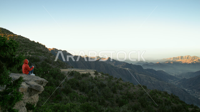شاب يجلس فوق صخرة اعلى قمة جبلية في مرتفعات جبال السودة في مدينة ابها في المملكة العربية السعودية، الاشجار والنباتات الخضراء، القمم والمرتفعات الجبلية، معالم عسير السياحية،