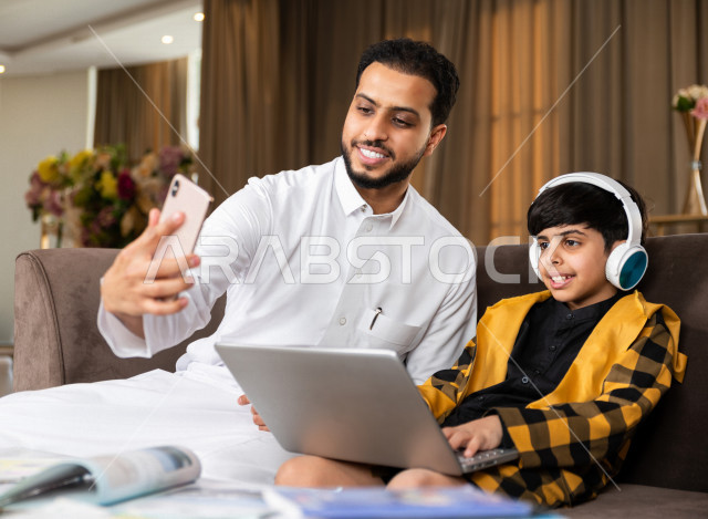 تلميذ سعودي خليجي يرتدي سماعة الراس و يستخدم الحاسوب المحمول ،  يدرس في المنزل مع والده ، التقاط صورة سيلفي اثناء الدراسة  ، التعلم عن بعد ، مفهوم التعليم عبر الانترنت ، التعليم المنزلي اثناء الحجر الصحي و تفشي فيروس كورونا ، العزلة الذاتية .