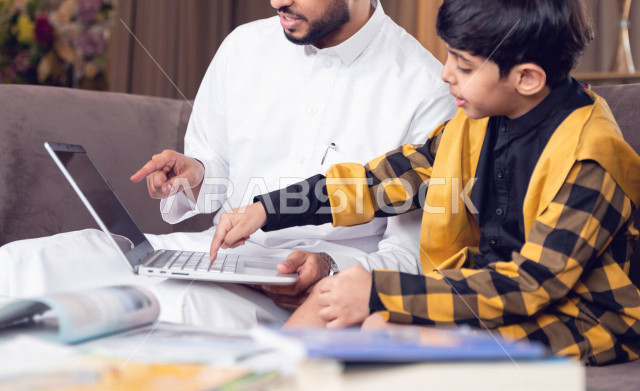 تلميذ سعودي خليجي يدرس في المنزل مع والده ،الذي يتبع معه حصص الاونلاين عن طريق الحاسوب المحمول  ، التعلم عن بعد ، مفهوم التعليم عبر الانترنت ، التعليم المنزلي اثناء الحجر الصحي و تفشي فيروس كورونا ، العزلة الذاتية .