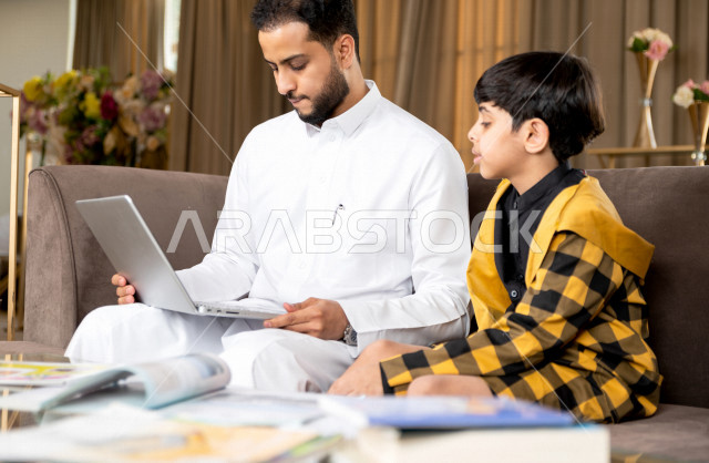 A Saudi Gulf student studying at home with his father, who follows with him online classes via a laptop, distance learning, the concept of online education, home education during quarantine and the outbreak of the Corona virus, self-isolation.