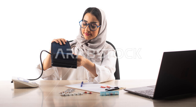 Using a blood pressure monitor, working in the medical sector, using a laptop computer, a portrait of a veiled Saudi Arabian Gulf doctor wearing a white medical coat and medical glasses, providing medical advice and services, white background