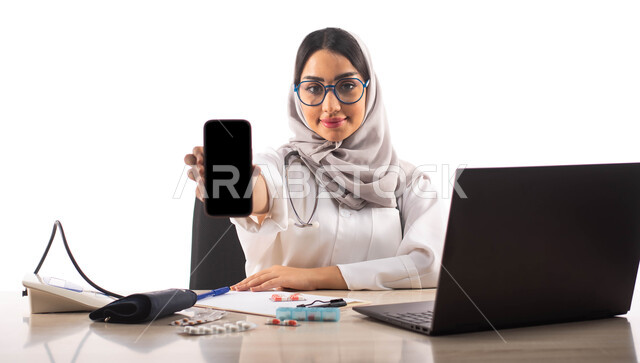جهاز قياس ضغط الدم, عرض شاشة سوداء فارغة عبر الهاتف المحمول, استخدام جهاز الحاسوب المحمول, البحث والتطوير ،  بورتريه لطبيبة عربية خليجية سعودية محجبة ترتدي المعطف الطبي الابيض والنظارات الطبية ,خلفية بيضاء