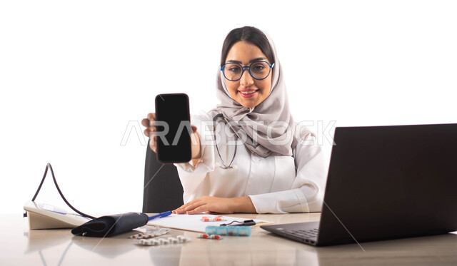  استخدام جهاز الحاسوب المحمول, جهاز قياس ضغط الدم, عرض شاشة سوداء فارغة عبر الهاتف المحمول, البحث والتطوير ،  بورتريه لطبيبة عربية خليجية سعودية محجبة ترتدي المعطف الطبي الابيض والنظارات الطبية ,خلفية بيضاء