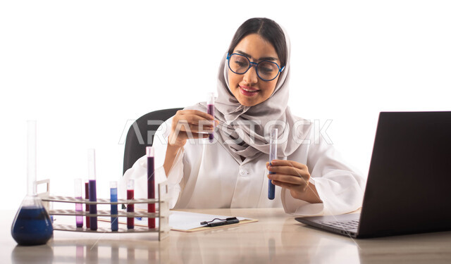 ايماءات تدل على الفرح والسعادة, الجلوس في معمل الكيمياء ، بورتريه لطبيبة مختبرات وتحاليل طبية عربية خليجية سعودية محجبة ترتدي المعطف الطبي الابيض و تحمل بيدها انبوب زجاجي , استخدام انابيب الاختبار لاجراء التجارب في المختبر ، التفاعلات الكيميائية ،البحث وا