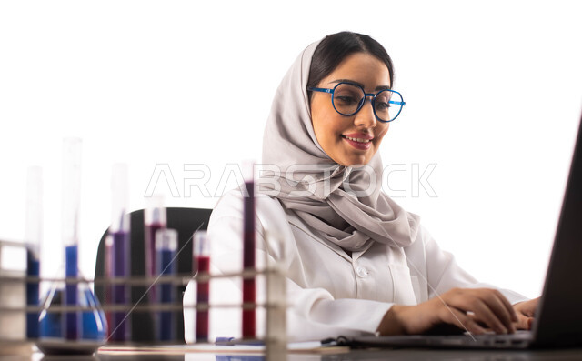 استخدام جهاز الحاسوب المحمول, البحث والتطوير ، الجلوس في معمل الكيمياء ، بورتريه لطبيبة مختبرات وتحاليل طبية عربية خليجية سعودية محجبة ترتدي المعطف الطبي الابيض والنظارات الطبية ,استخدام انابيب الاختبار لاجراء التجارب في المختبر ، التفاعلات الكيميائية ، م