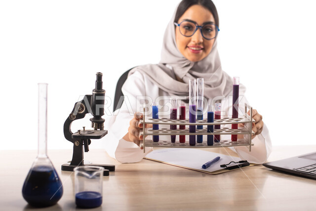 التفاعلات الكيميائية ، الجلوس في معمل الكيمياء ، بورتريه لطبيبة مختبرات وتحاليل طبية عربية خليجية سعودية محجبة ترتدي المعطف الطبي الابيض والنظارات الطبية وتحمل بيدها انابيب الاختبار لاجراء التجارب في المختبر ، البحث والتطوير ، مفهوم الكيمياء الحيوية, خلفي