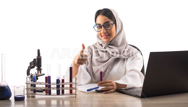 Gestures indicating the mark of quality, sitting in the chemistry laboratory, a portrait of a veiled Saudi Arabian Gulf doctor wearing a white medical coat and medical glasses, using a laptop, using test tubes to conduct experiments in the laboratory, che