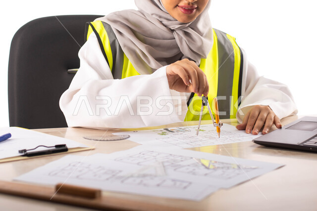 ارتداء بدلة السلامة , استخدام الفرجار الهندسي و الأدوات والمعدات على مخطط تصميم هندسي ،  بورتريه لمهندسة معمارية عربية خليجية سعودية محترفه ومبتسمة داخل مكتب هندسي ، خلفية بيضاء