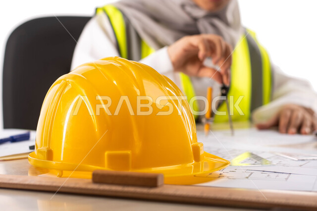 مهن ووظائف ، استخدام أدوات ومعدات هندسية على مخطط تصميم هندسي ، ارتداء بدلة السلامة, بورتريه لمهندسة معمارية عربية خليجية سعودية محترفه داخل مكتب هندسي  خوذة الوقاية الصفراء ، خلفية بيضاء