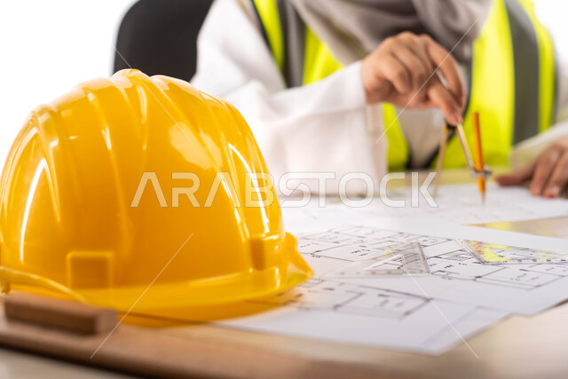  خوذة الوقاية الصفراء، استخدام أدوات ومعدات هندسية على مخطط تصميم هندسي ، ارتداء بدلة السلامة, بورتريه لمهندسة معمارية عربية خليجية سعودية محترفه داخل مكتب هندسي ، مهن ووظائف ، خلفية بيضاء
