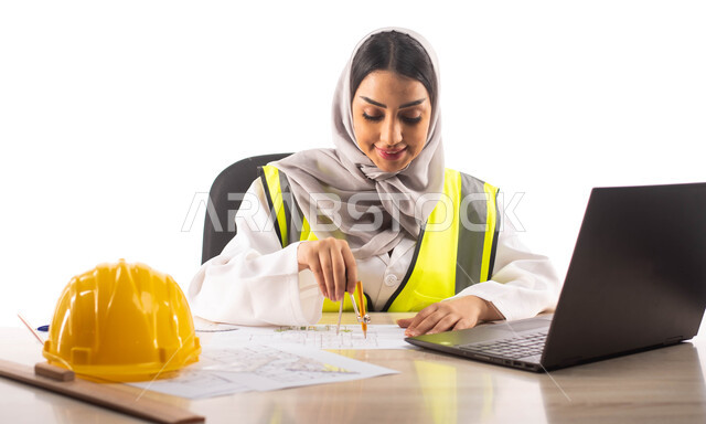  ايماءات تدل على السعادة, استخدام أدوات ومعدات هندسية على مخطط تصميم هندسي ، ارتداء بدلة السلامة, بورتريه لمهندسة معمارية عربية خليجية سعودية محترفه داخل مكتب هندسي ، إستخدام جهاز الحاسوب المحمول للإنجاز وتتبع المهام ، خوذة الوقاية ،مهن ووظائف ، خلفية بيض