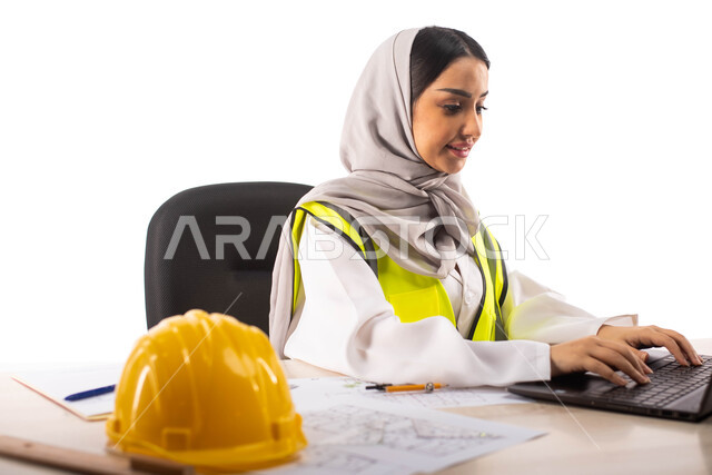 ايماءات تدل على الفرح, إستخدام جهاز الحاسوب المحمول للإنجاز وتتبع المهام ، استخدام أدوات ومعدات هندسية على مخطط تصميم هندسي ، ارتداء بدلة السلامة, بورتريه لمهندسة معمارية عربية خليجية سعودية محترفه داخل مكتب هندسي ، مهن ووظائف ،خوذة الوقاية الصفراء ،خلفية