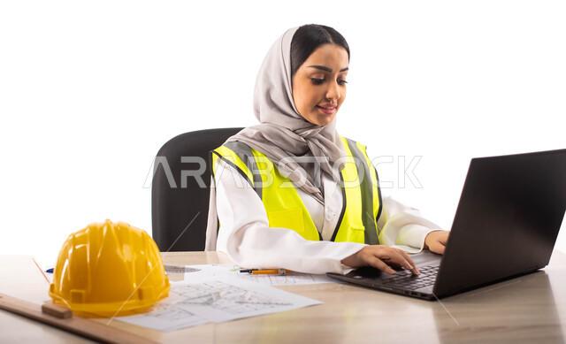 إستخدام جهاز الحاسوب المحمول للإنجاز وتتبع المهام ، ايماءات تدل على الفرح,  استخدام أدوات ومعدات هندسية على مخطط تصميم هندسي ، ارتداء بدلة السلامة, بورتريه لمهندسة معمارية عربية خليجية سعودية محترفه داخل مكتب هندسي ، مهن ووظائف ،خوذة الوقاية الصفراء ،خلفي