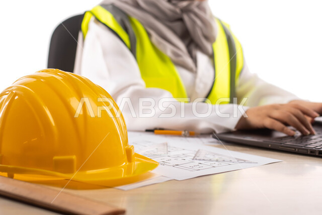 مهن ووظائف  ، استخدام أدوات ومعدات هندسية على مخطط تصميم هندسي ، ارتداء بدلة السلامة, بورتريه لمهندسة معمارية عربية خليجية سعودية محترفه داخل مكتب هندسي ، إستخدام جهاز الحاسوب المحمول للإنجاز وتتبع المهام ، خوذة الوقاية الصفراء ،خلفية بيضاء