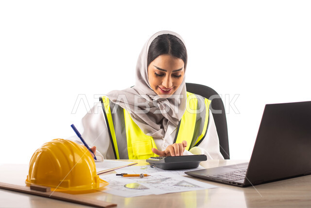  التخطيط لمشروع جديد, استخدام الآلة الحاسبة لإجراء العمليات الحسابية، ارتداء بدلة السلامة, بورتريه لمهندسة معمارية عربية خليجية سعودية محترفه داخل مكتب هندسي ، إستخدام جهاز الحاسوب المحمول للإنجاز وتتبع المهام ، خوذة الوقاية الصفراء ،مهن ووظائف ، خلفية بي