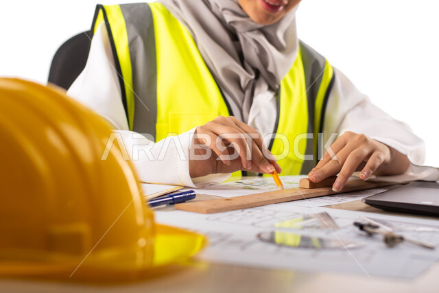 ارتداء بدلة السلامة, استخدام أدوات ومعدات هندسية على مخطط تصميم هندسي ،  بورتريه لمهندسة معمارية عربية خليجية سعودية محترفه داخل مكتب هندسي ، إستخدام جهاز الحاسوب المحمول للإنجاز وتتبع المهام ، خوذة الوقاية ،مهن ووظائف ، خلفية بيضاء