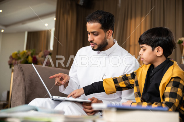 A Saudi Gulf student studying at home with his father, who follows with him online classes via a laptop, distance learning, the concept of online education, home education during quarantine and the outbreak of the Corona virus, self-isolation.